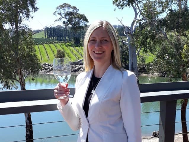 Blonde girl holding a glass of wine in front of a lake and vineyard