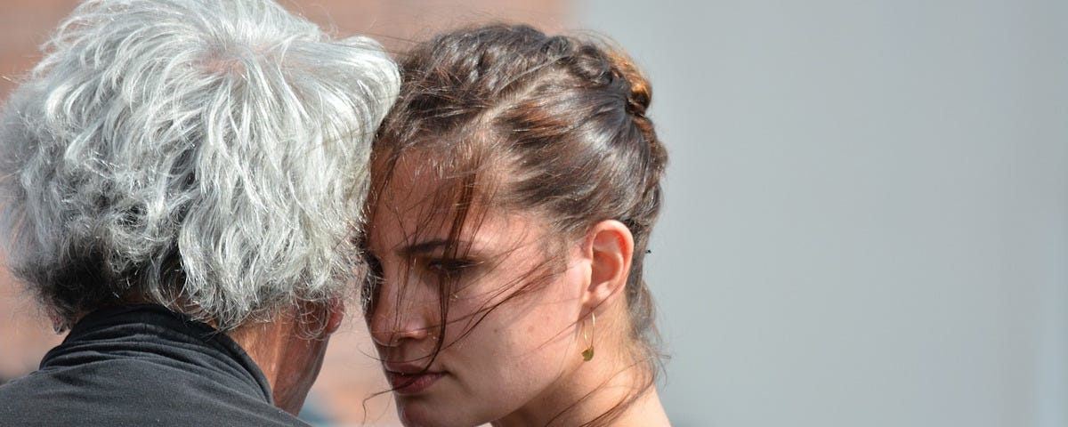 A beautiful young woman with her hair up in an elegant bun is dancing a tango with a much older man with shaggy grey hair. Her right temple is against his left, and she is gazing intently past him. A few strands of the woman’s hair have come loose and flown forward, clinging to her face and neck.