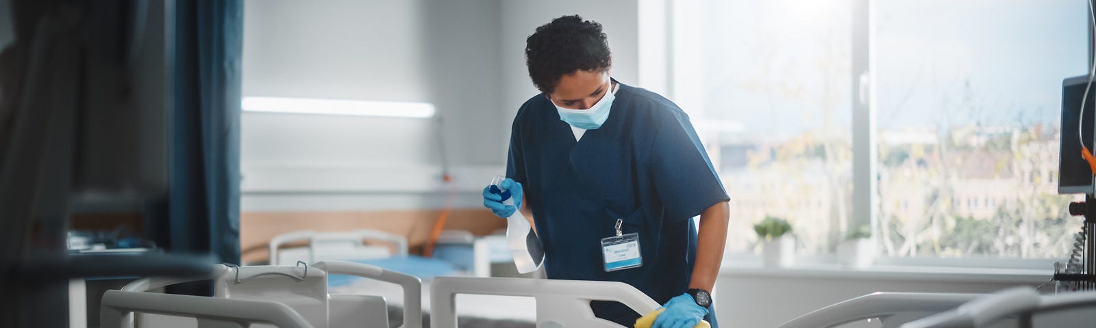 A masked and gloved health care worker disinfects a hospital bed.