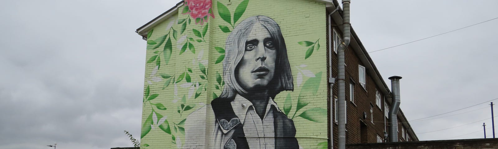 Large mural on a wall of the side of a building on an housing estate depicting a portrait of guitarist Mick Ronson, head and upper body, surrounded by flowers and greenery, also showing children on a park’s running track.