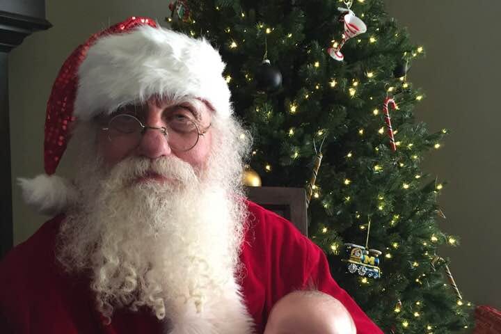 Bearded man with round gold frames sits in front of a Christmas tree in a Santa suit with a baby on his lap.