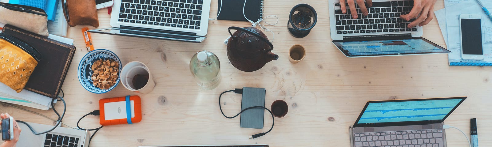 Multiple people collaborate at a table with laptops, phones, notebooks, pens, and coffee