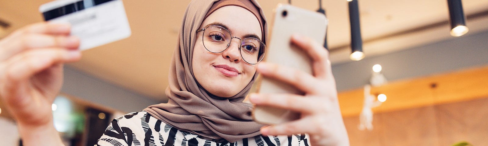 Woman with glasses in a hijab and black and white pattered short, looking at her phone in one hand and holding a credit card in another hand, with a slight smirk on her face.