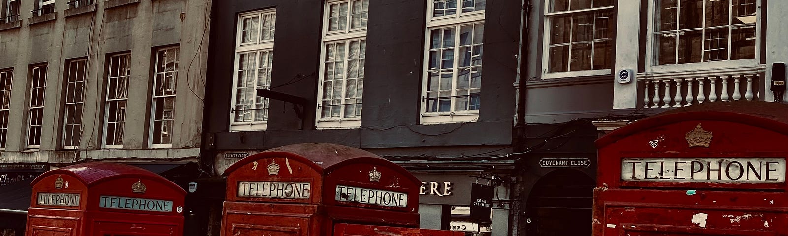 Three weathered, red, British telephone boxes stand upon the sidewalk in front of a row of five-story granite buildings.