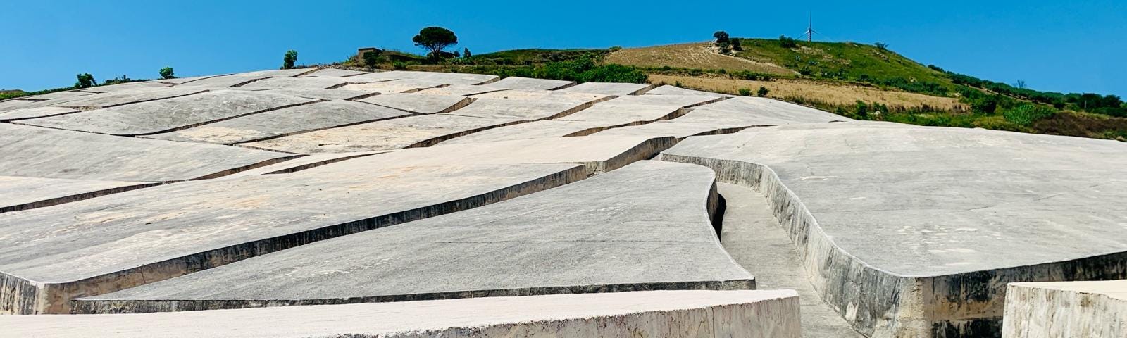 Large flat-top concrete structure climbs up a hillside with the top of the hill visible in the distance. There are pathways carved out of the concrete.