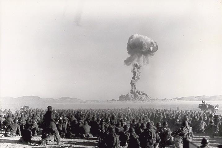 A black and white photo shows military troops in the foreground and in the background a mushroom cloud rises from a nuclear test in the Nevada desert.