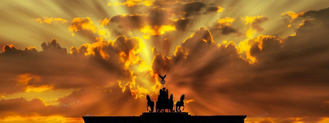 A silhouette of the Brandenburger Tor (Gate) in Berlin, Germany against the golden rays of sunrise behind clouds.