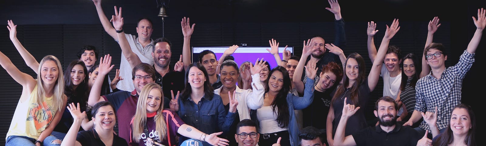 Foto com pessoas de mão levantadas e de frente e no fundo uma TV ligada. Foi durante o Open House da olist em 2019