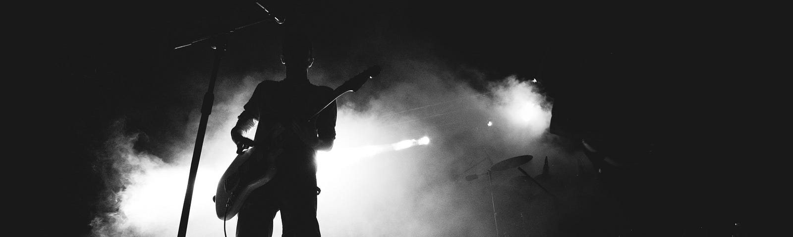 A man in silhouette on a stage plays the guitar. He is outlined by the lights behind him. It is very dramatic.