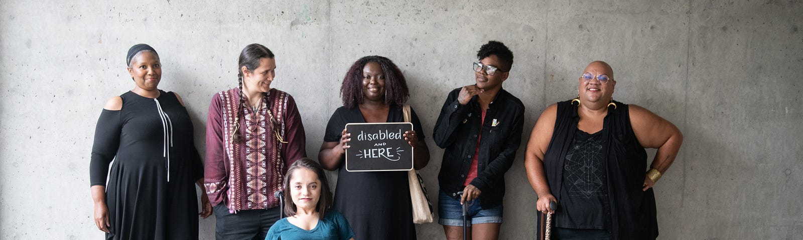 Six disabled people of colour smile and pose in front of a concrete wall.
