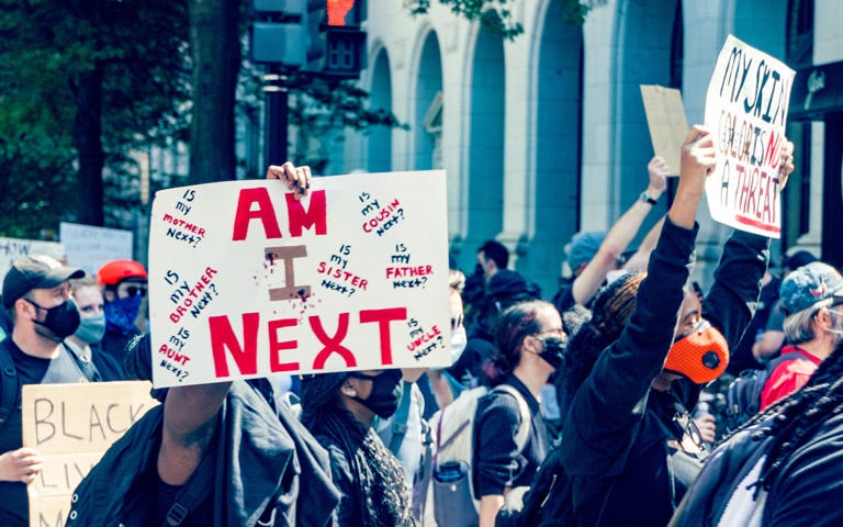 Demonstrators, most of whom are wearing face masks, stand or walk in front of a pedestrian crossing signal. They are facing something that is outside of the frame to the right. Several demonstrators are holding signs. The sign facing the camera has mostly red text and reads, “Am I next?”