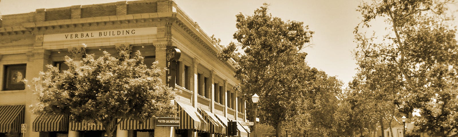 A street corner in Hunter Mudd College’s campus in Claremont, California. The “Verbal Building” is visible, as are pedestrians, pparked cars, and trees along the streeet.