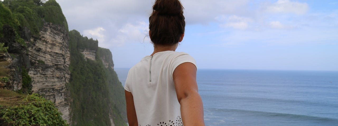 A woman standing at a cliff’s edge overlooking the ocean with her hand being held by another.