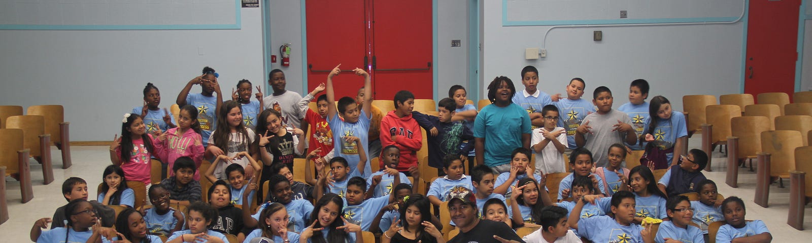 Two men crouch in front of a group of children seated in a school auditorium