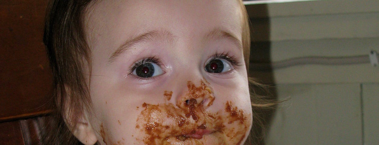 A close-up of a young brown-eyed girl looking at the camera with brown smears spread over her lower face, hand, forearm and bib.