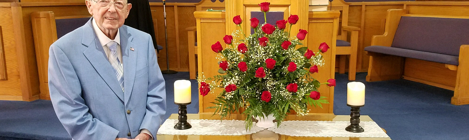 80 year old man in sport coat, dress pants, and neck tie standing beside a flower arrangement of roses on a table at the front of a church. The stage is behind him and you can see the pulpit.