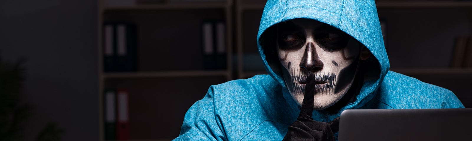 A person with skeleton face paint in a hoodie in a dark library behind a laptop, with their finger in a “shh” position.