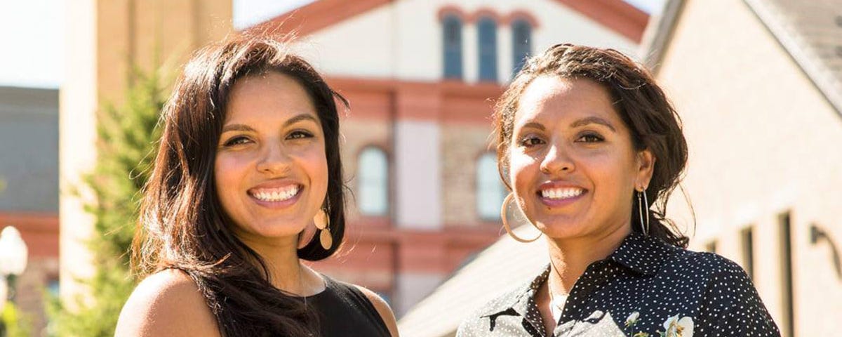 Diana and Denise Lopez stand side-by-side, smiling on the Regis Northwest Denver Campus with Main Hall and the Chapel in the background