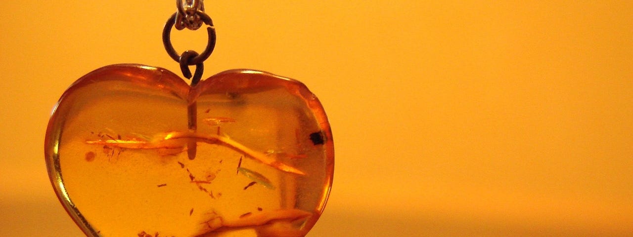 An amber heart-shaped pendant hanging in front of a similarly covered wall. There appears to be flecks of gold swimming in the pendant.