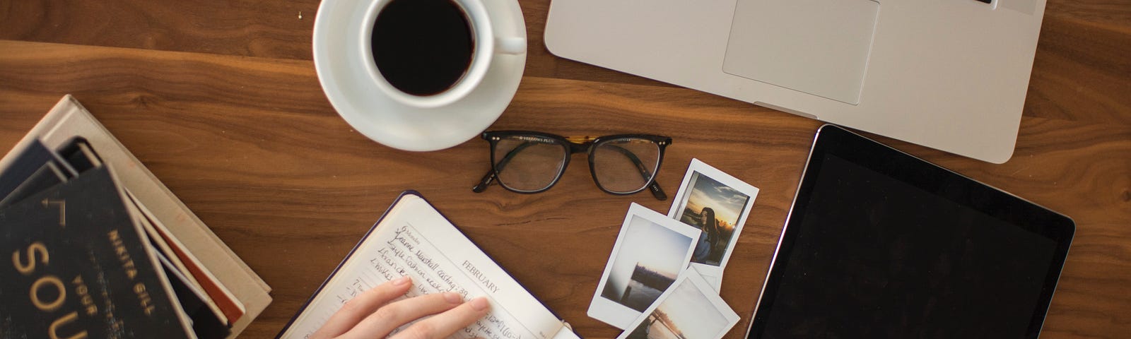 A person is writing something down in a notebook on a wooden table. There are books, 3 photos, a laptop, a pair of glasses, and a cup of coffee.