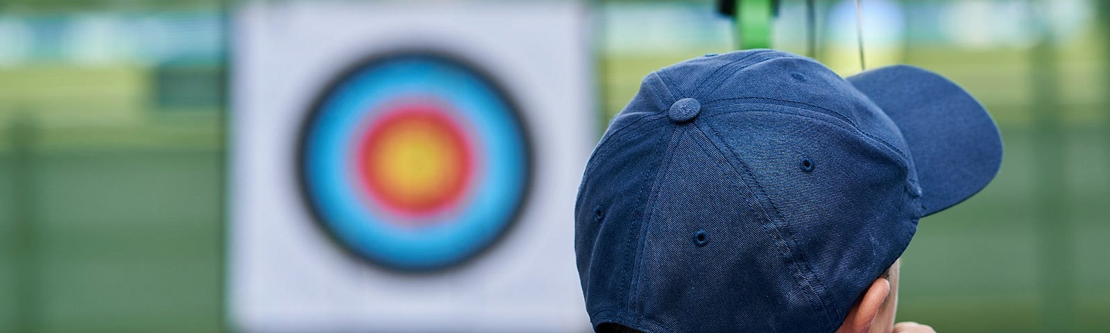 Boy wearing blue cap, blue armband, and white T-shirt aiming his bow and arrow at a target in the background.