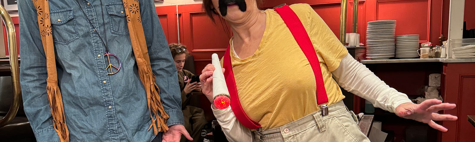 Woman in red suspenders wears a mustache. Young man, her son, to the left, looks on.