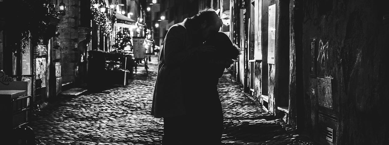 Black and white image of a couple kissing in a dark street.