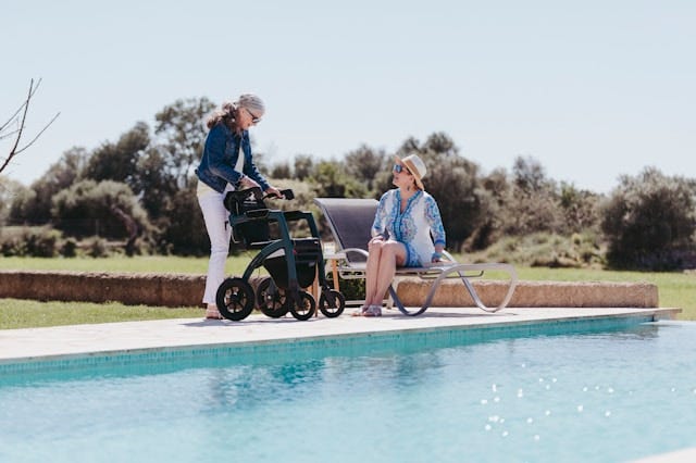 Two older women, one of them standing behind a a walker  by a swimming pool. The other woman is seated.