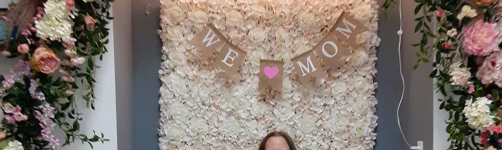 I’m seated in front of backdrop of closely-packed white roses with a burlap banner reading “WE (picture of heart) MOM.” The white-roses backdrop is set on a slate-blue inset in the wall. The white wall that frames the floral inset is covered from top to bottom with a graceful swag of white and pink flowers and greenery. I’m a white woman wearing glasses, a caramel-colored t-shirt, and a skirt with a muted abstract pattern. I’m holding a small jar of colorful flowers and greenery.