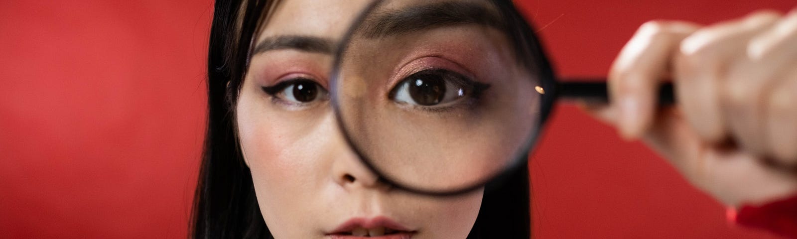 Picture of a woman with shoulder length dark hair standing in front of a red background. She is looking at you through a magnifying glass.