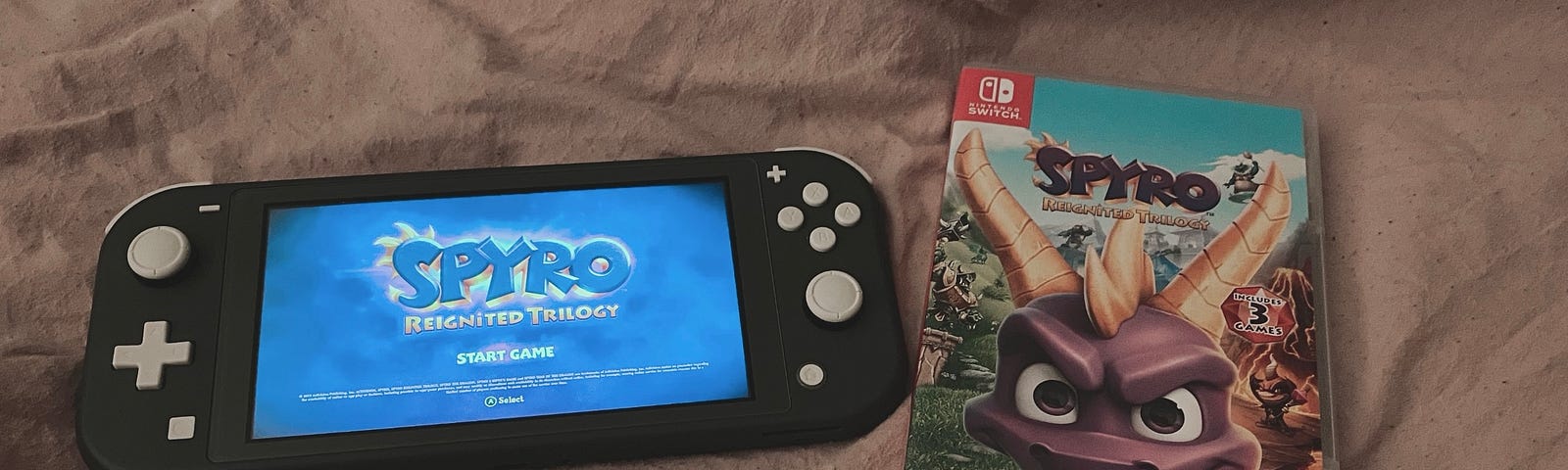A gray Nintendo switch lite with the Spyro the Dragon: Reignited Trilogy home screen on the left. Next to the switch is the case for the Spyro game. Both lying flat on a pink blanket.