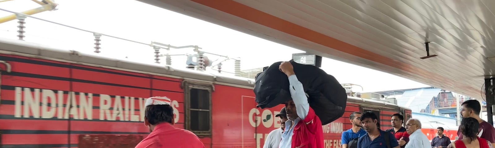 Railway platform with a train that has arrived. The train says “Indian Railways.” C couple of coolies/porters are loading luggage. Other people are wheeling their suitcases.