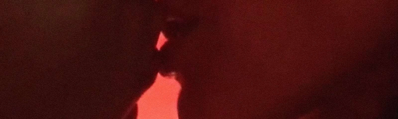Dos personas se acercan para besarse en primer plano, sus labios se rozan; las siluetas dejan ver luz roja al fondo.