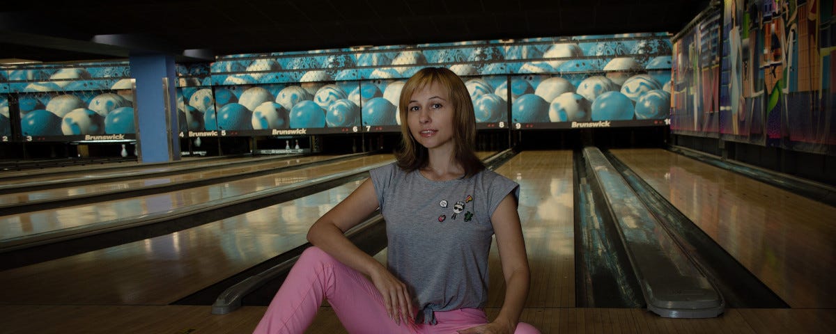 An attractive, young, blonde woman wearing a grey t-shirt and pink jeans, sitting on the floor in front of a ten-pin bowling lane, with a bowling ball in her hand.