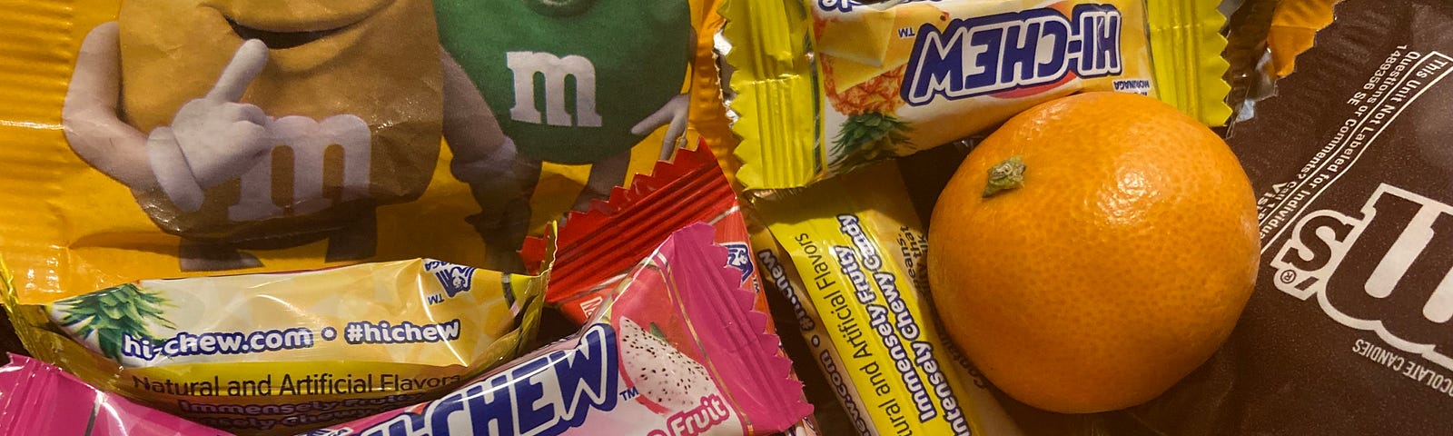 An assortment of fun-size original and peanut M&M’s in brown and yellow packets respectively, three Mandarin oranges, one pink Hi-Chew dragon fruit candy, and three yellow Hi-Chew pineapple candy.
