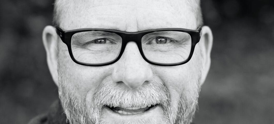 Black and white picture of an older man who is bald and wearing glasses and has a beard with gray hair.