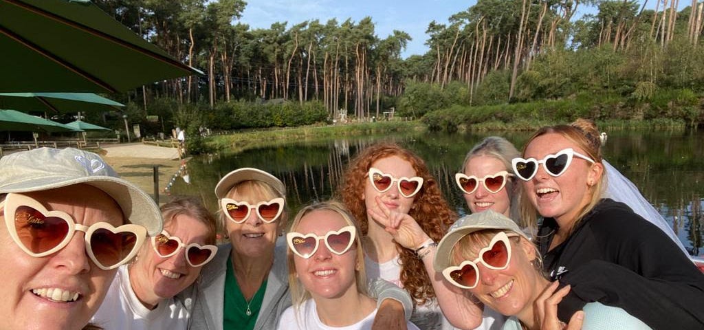 A group of women in love heart sunglasses, standing in front of a lake.