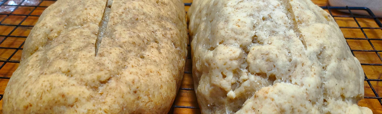 Two loaves of homemade bread cooling on a wire rack. The loaf on the right is a lighter color than the loaf on the left, which is a tan color. The rack is sitting on a wooden cutting board.