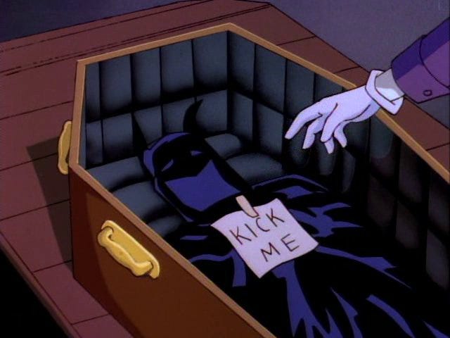 A screenshot from ‘Batman: The Animated Series’. The Joker’s hand can be seen attaching a “kick me” sign to Batman’s cape and cowl, inside a coffin.