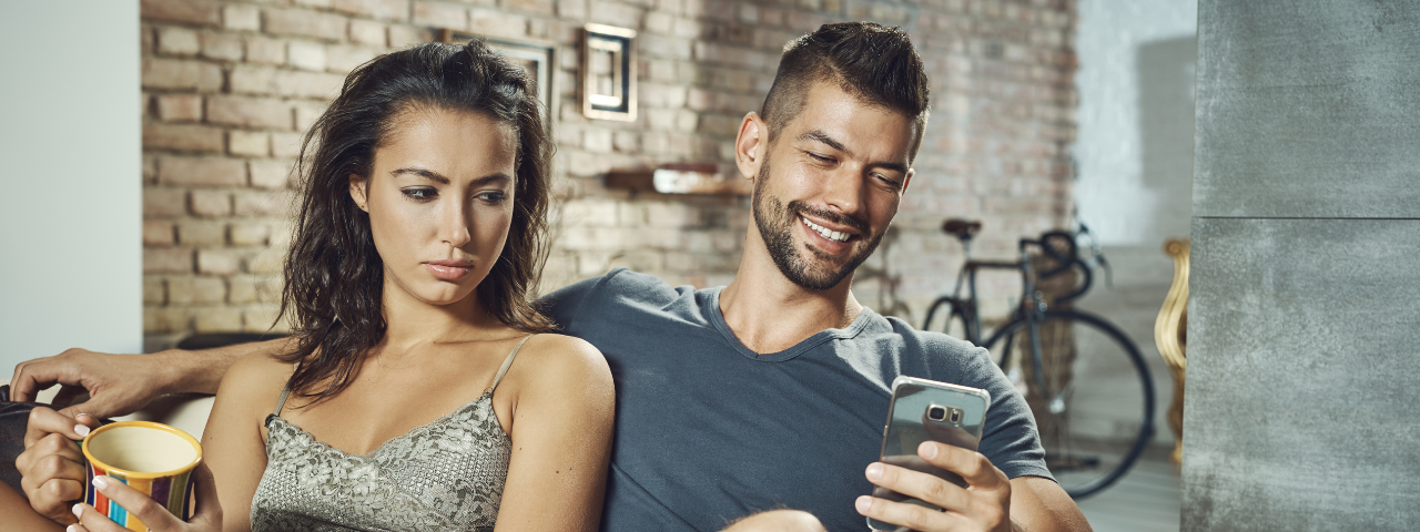 A woman looking jealous as her boyfriend smiles at a phone — How to not let jealousy ruin your relationship