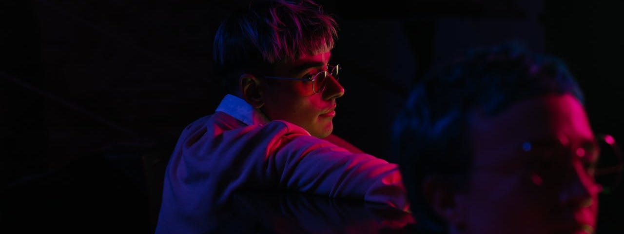 Two people sit in the dark with only a bit of purple light illuminating them. One person looks over their shoulder toward the back of the other person.