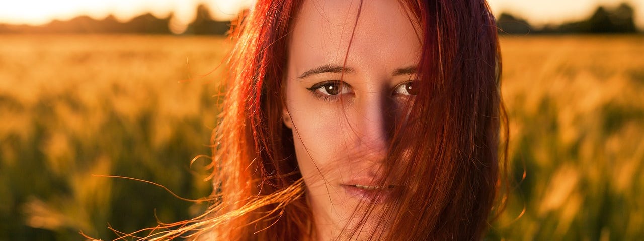 A woman with long, red hair and a sad expression standing in front of a wheat field.