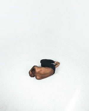 Muscular man curling himself into a fetal position in a pure white room. He is topless and only wearing black pants.