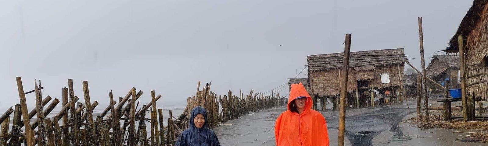 Two intrepid researchers walking through a small fishing village, with thatch huts and wooden fencing, in a torrential downpour.