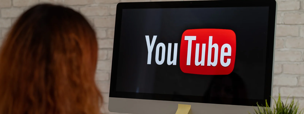 This image shows a woman viewing the YouTube logo on a large desktop computer screen. The setting appears to be a home office with a white brick wall in the background and a small potted plant on the desk.