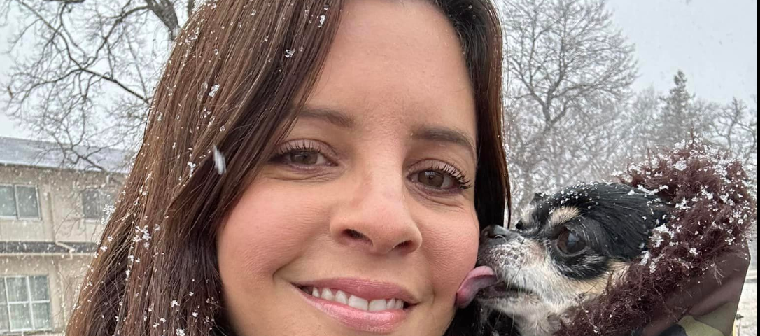 Woman standing in the snow getting licked on the face by a chihuahua wearing a fur collared coat