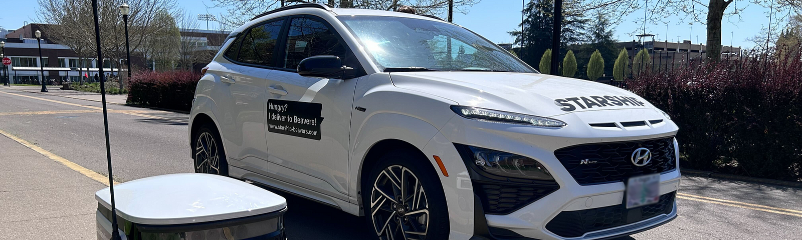 Image of a Starship Robot next to a white car with decals that read “Starship” and “Hungry? I deliver to Badgers” to make the white car look like a Starship Delivery Robot