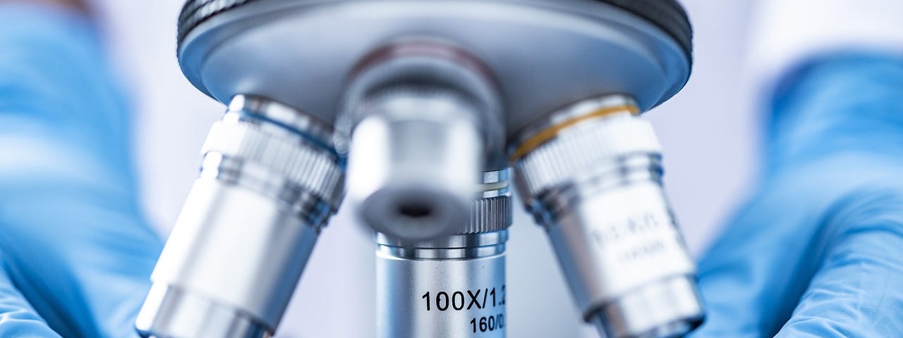 Image shows two hands, one on each side of the image, wearing blue latex gloves and in the middle of the image, the parts of a microscope which enlarges whatever lies under it.