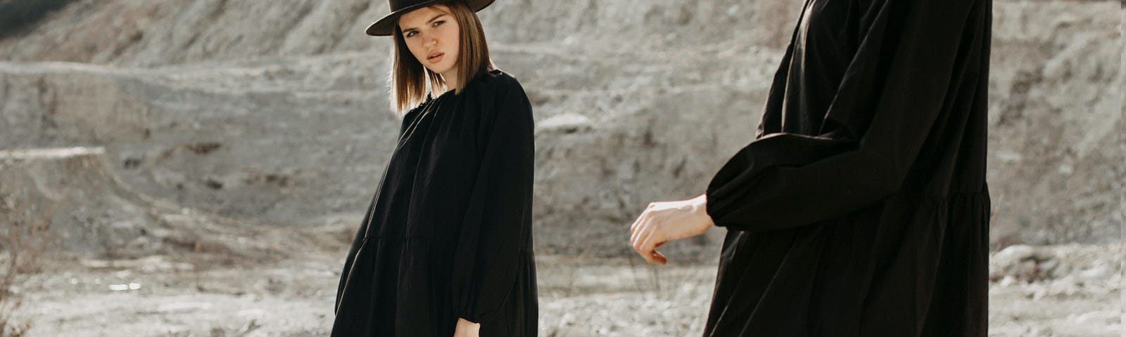 A woman dressed in black in a rocky terrain stares at a mirror/twin version of herself