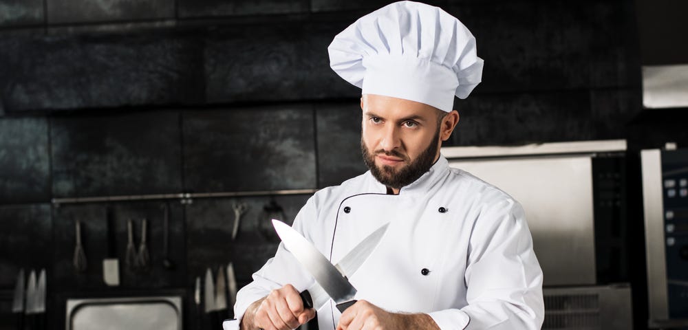 Chef in whites standing in a professional kitchen holding crossed knives